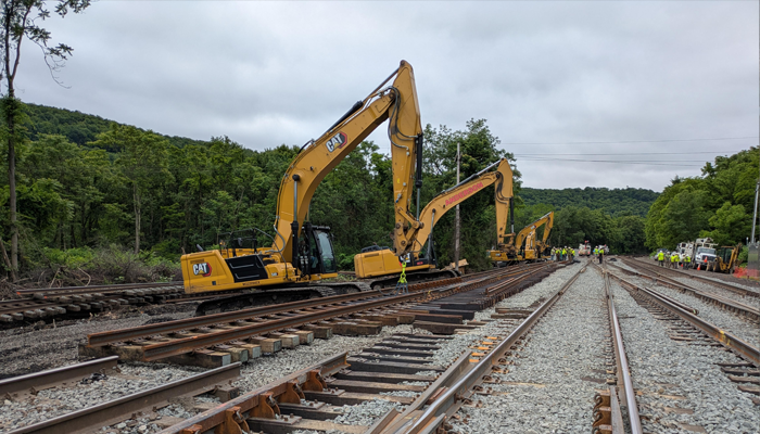 Two CAT excavators working on railroad track installation at a multi-track rail interlocking, with a construction crew visible in the background
