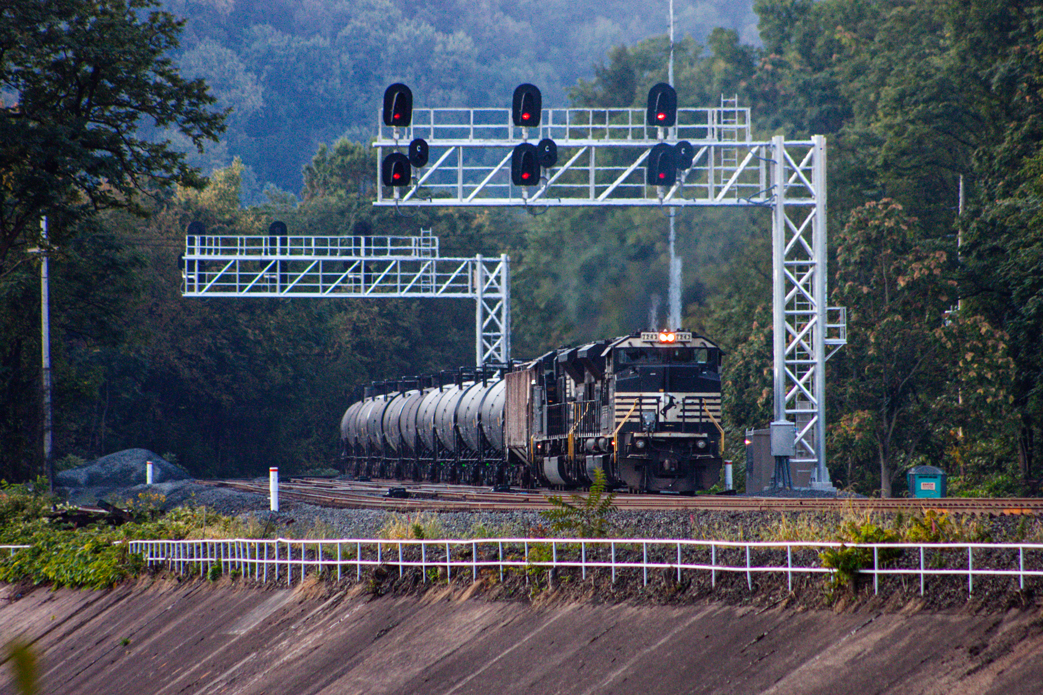 A Norfolk Southern freight train pulling tank cars passes beneath a signal bridge on a multi-track rail corridor surrounded by wooded hills.