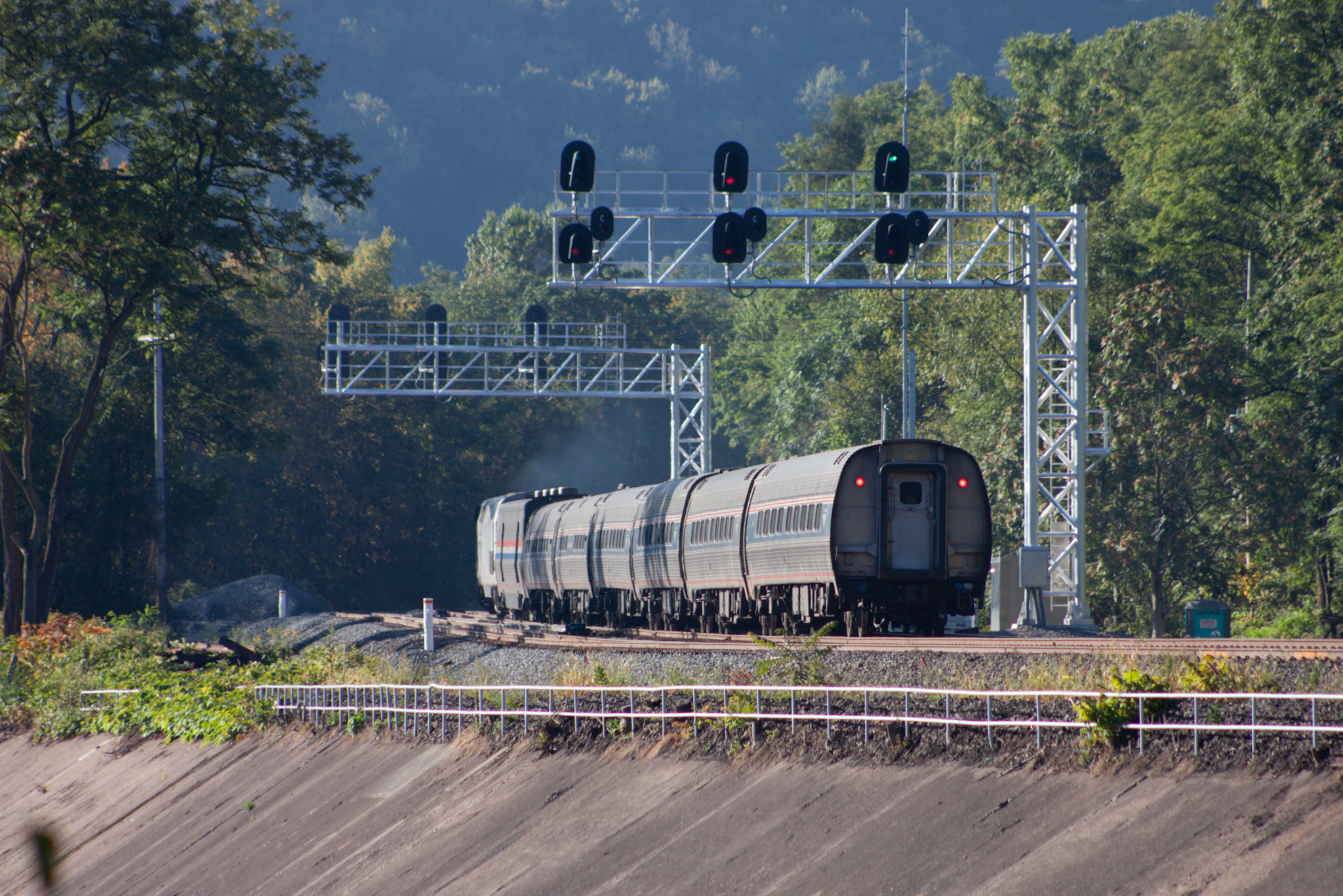 The rear of an Amtrak passenger train passing beneath a signal bridge on a rail corridor surrounded by wooded hills.